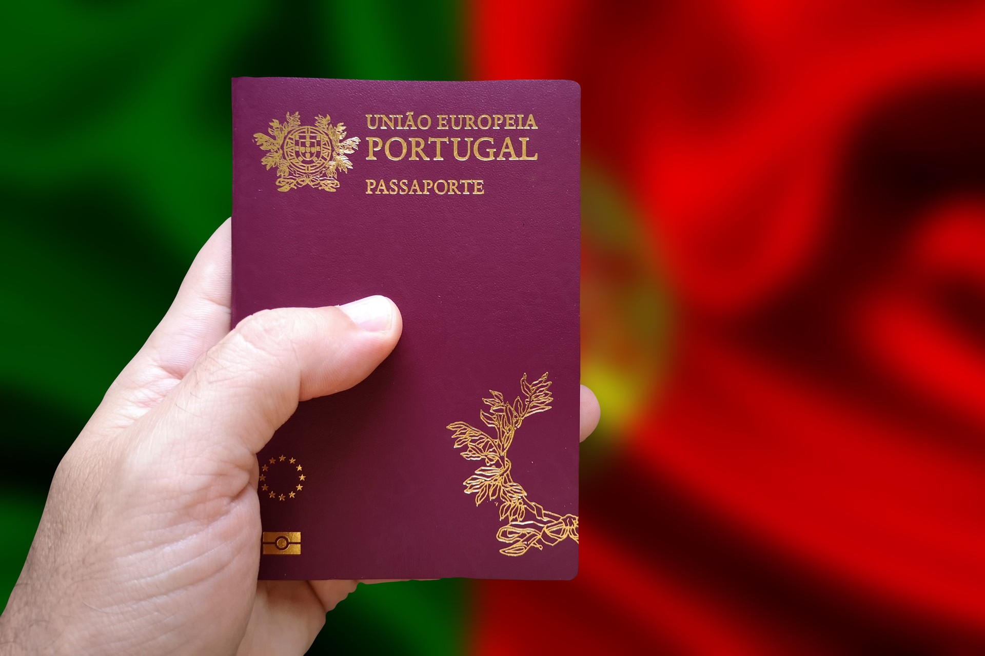 Man's hand holding Portuguese passport with defocused Portuguese flag in the background. Written in Portuguese: European Union Portugal. Passport.